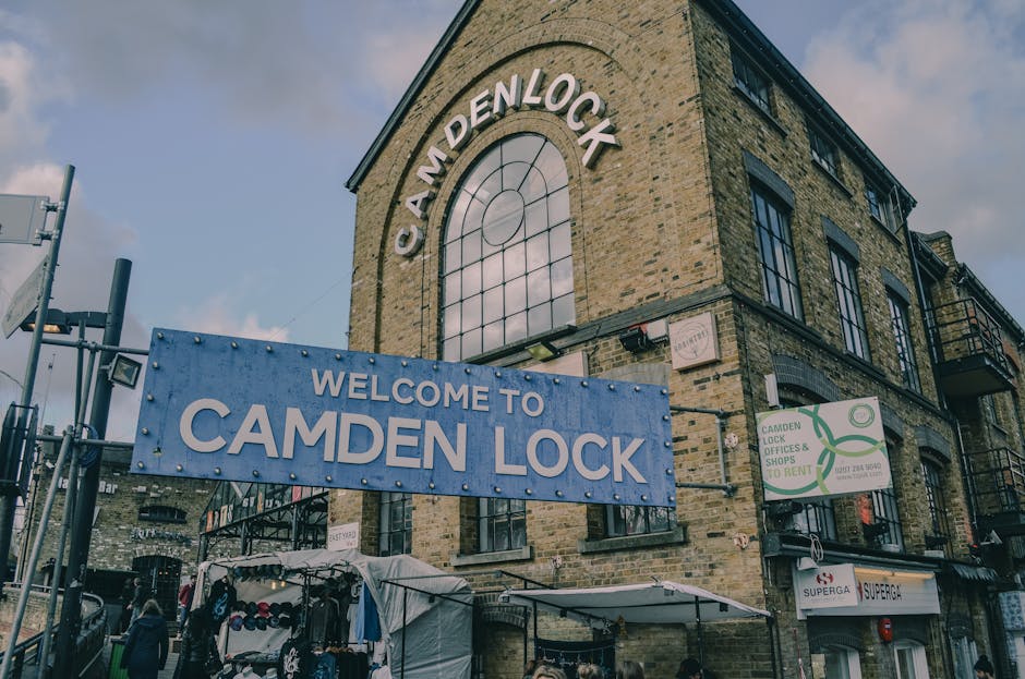 A three-story brick building with large, arched windows and black metal balcony railings on the upper floors, situated in an urban setting under a partly cloudy sky. A prominent blue sign reading 'Wel