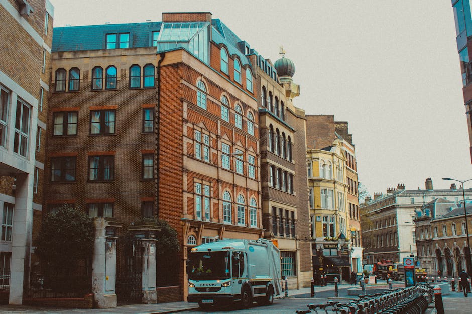 A city street scene showing a variety of multi-story buildings constructed with brick, stone, and ornate architectural details. In the foreground, a light blue waste collection vehicle is parked on th