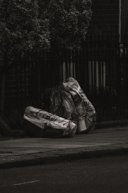 Two large black garbage bags filled with household waste are positioned on a paved sidewalk next to a curb. The bags appear to be made of thick plastic, with some creases and folds visible on their su