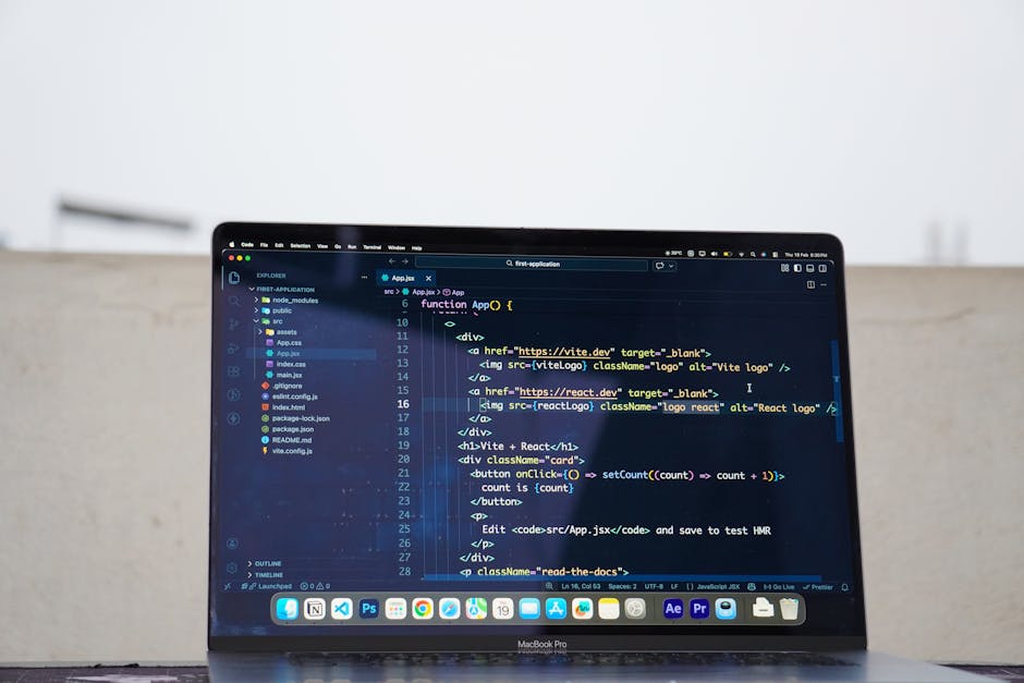 A person with dark hair, wearing a dark jacket, is seated indoors at a desk and is viewed from behind, working on a dual-monitor computer setup. The primary monitor displays lines of programming code 